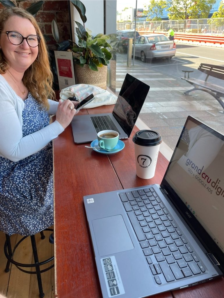 Casey smiling with 2 laptops and 2 coffees at 7 Origins Malop