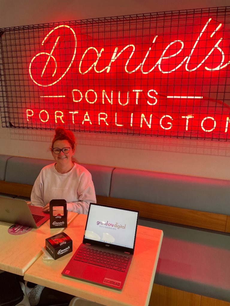 Casey smiling with 2 laptops and 2 donuts at Daniels Donuts